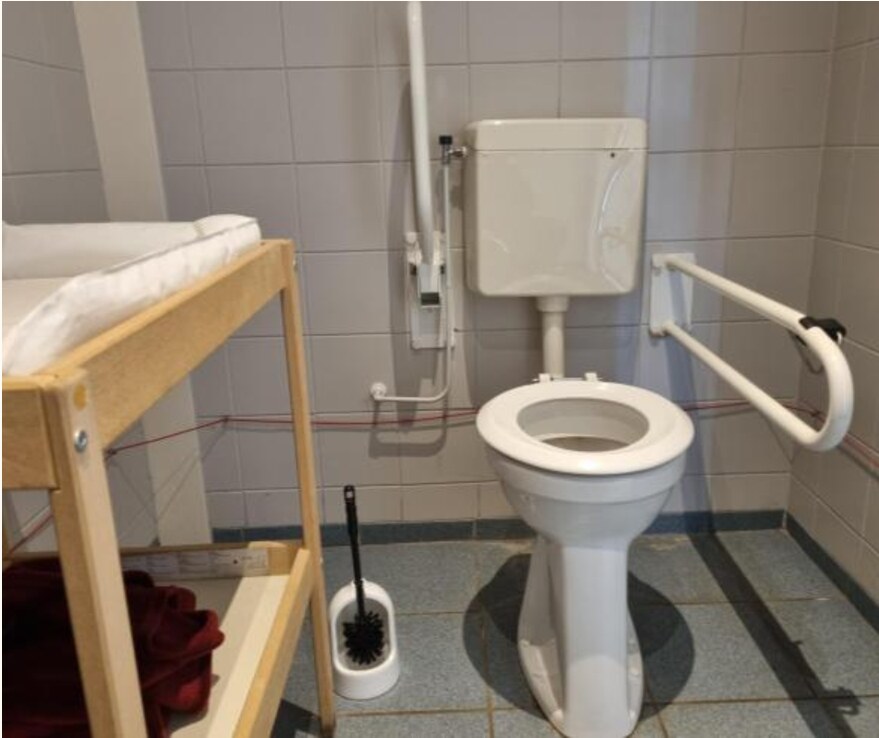 Foto van een toilet met aan beide kanten een opklapbaar toiletbeugel, en een verschoontafel er schuin voor. Er is geen ruimte voor een rolstoel om als persoon zelfstandig naar de WC te verplaatsen.
