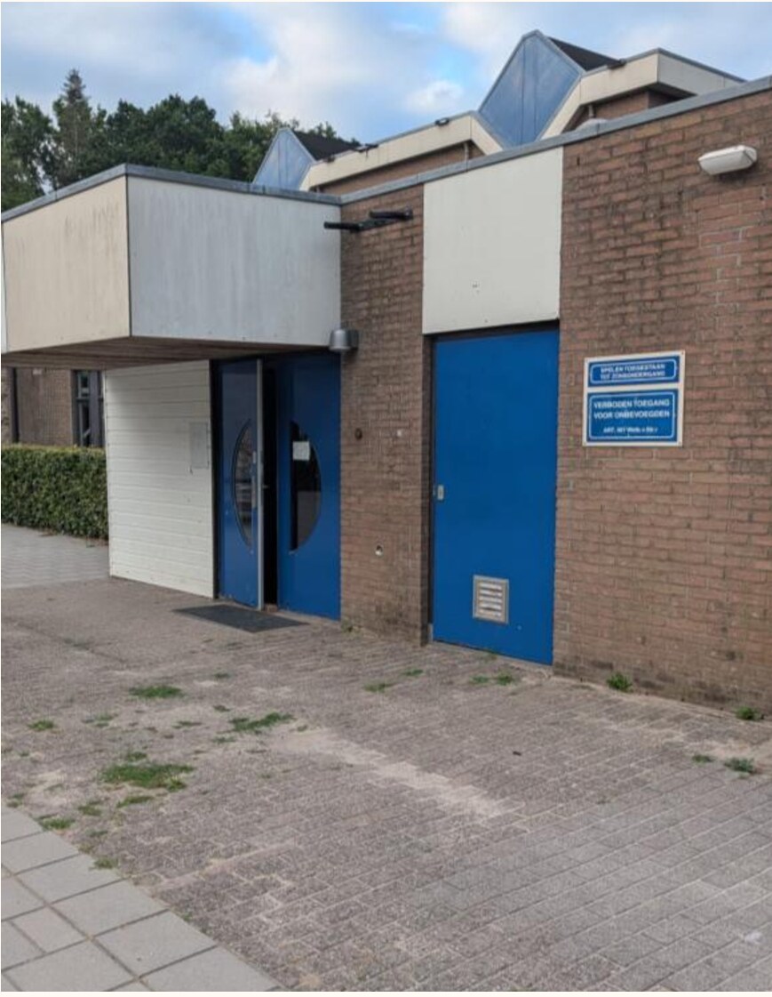 Foto van de hoofdingang van een zaalsportaccommodatie. De hoofdingang laat twee (niet automatische) klapdeuren zien, waarvan de rechterdeur vastzit.