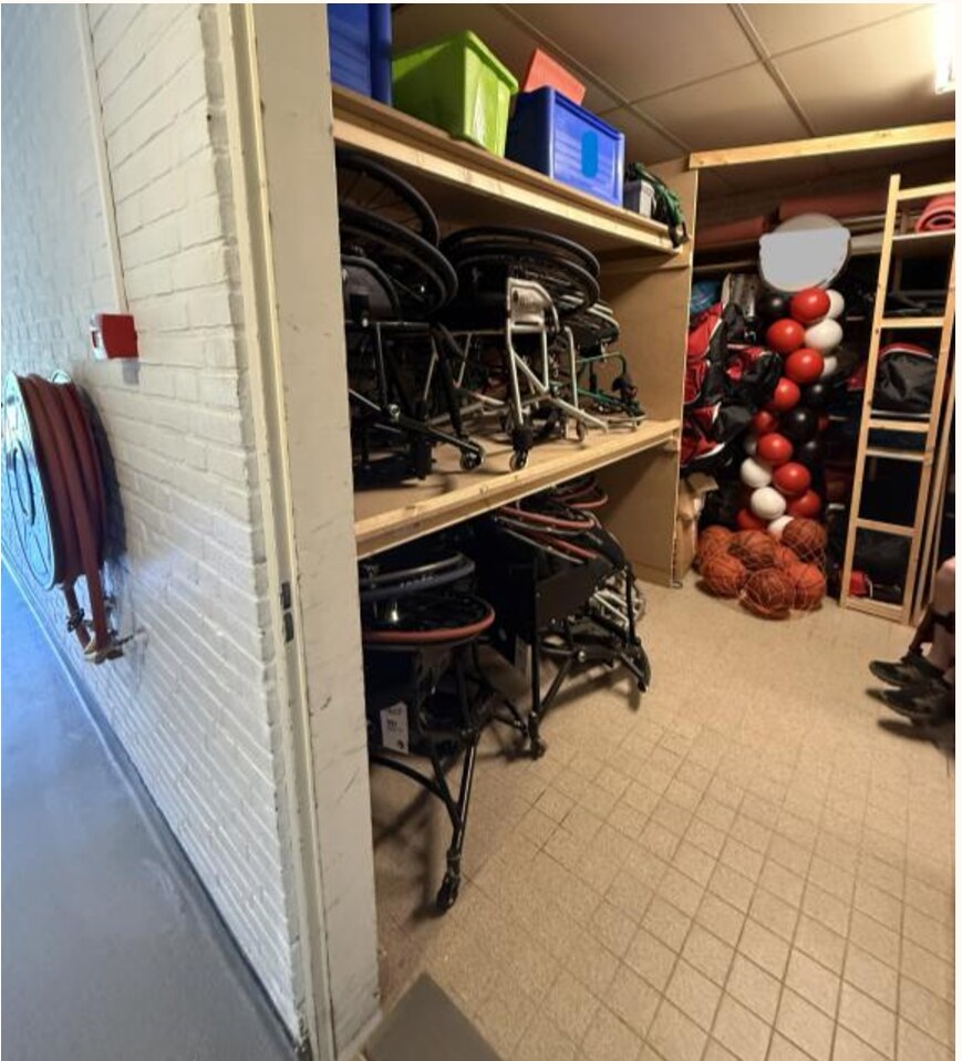 Foto van een deel van een opslagruimte. Links van de deuropening zijn op twee verschillende hoogtes een paar sportrolstoelen opgeborgen.