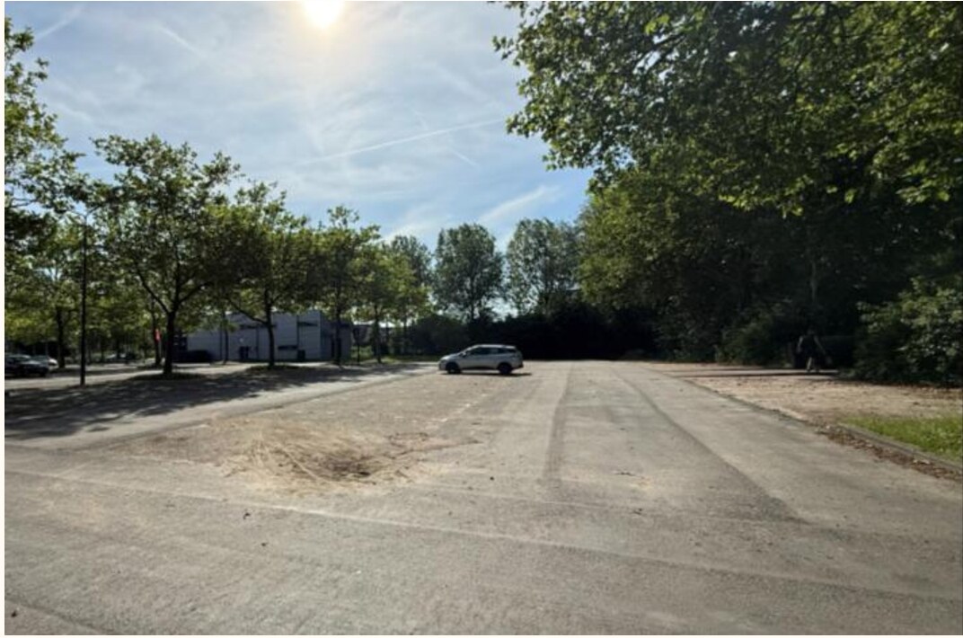 Foto van een deel van de parkeerplaatsen bij een zaalsportaccommodatie. De parkeerplaatsen zijn niet belijnd en er is veel open ruimte.