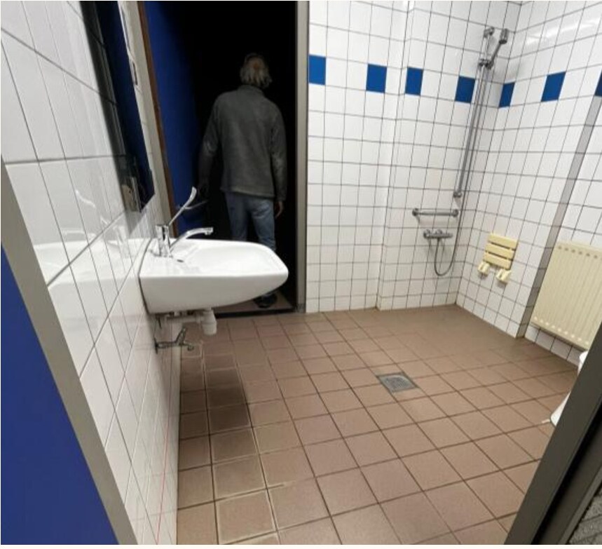 Foto van een aangepaste doucheruimte voor één persoon, te betreden via twee kleedkamers. Wastafel is op zithoogte, evenals bedieningsknoppen van de douche. Behalve de douchekop, die op stahoogte hangt.